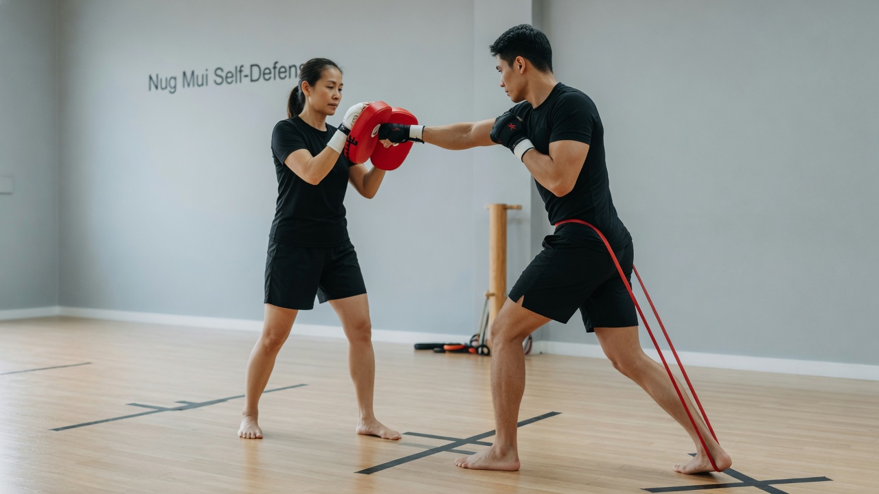 A clean, well-lit martial arts studio where a coach holds a red focus mitt